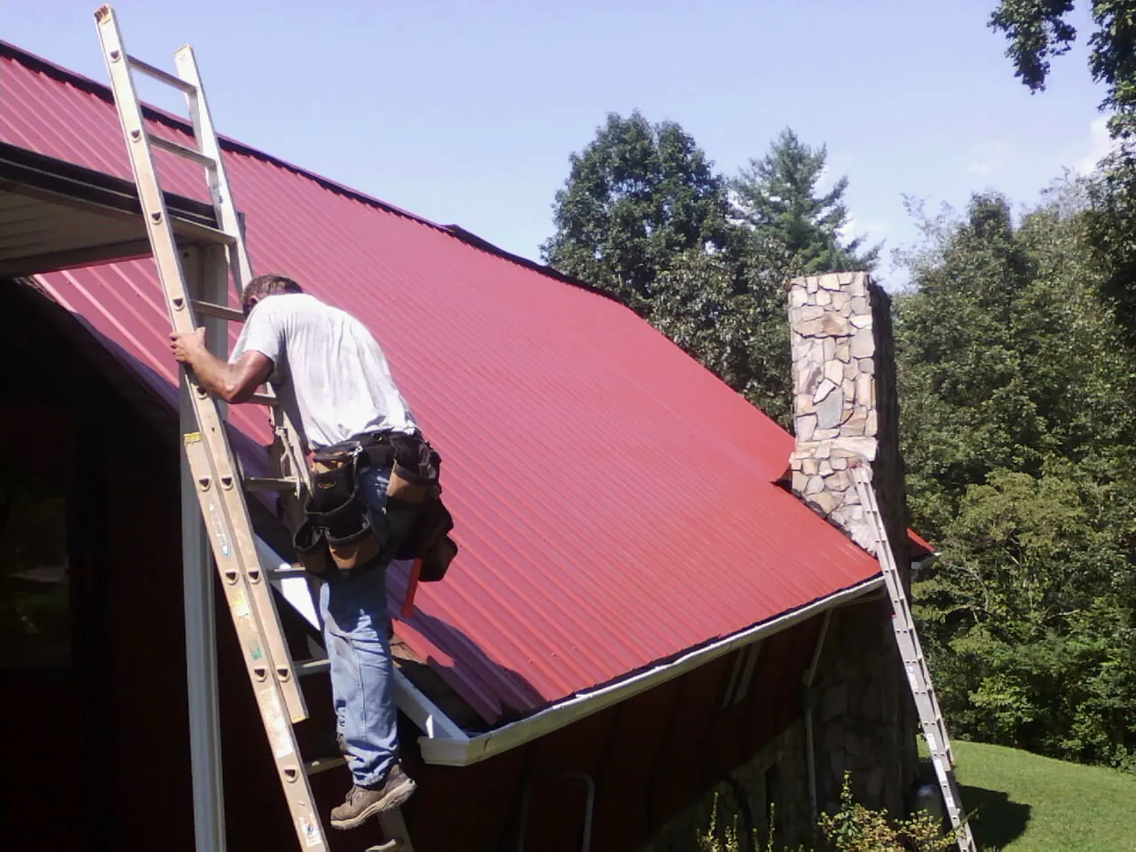 Roofer on a ladder beside a red metal roof and stone chimney — Soffit & Fascia Repair by Roofing Brothers in Gladewater